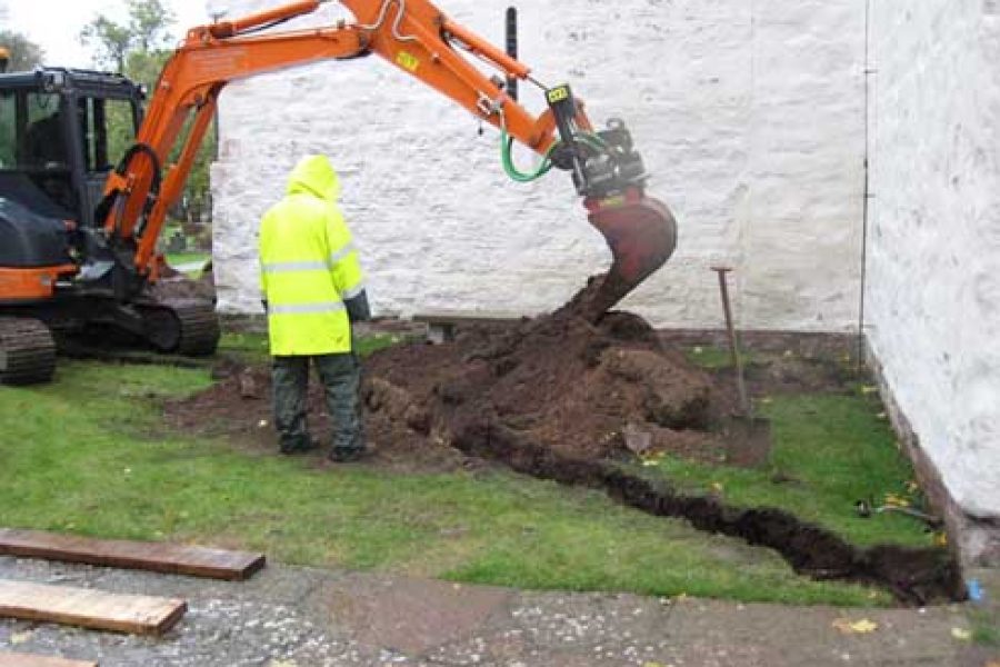 En grävmaskin gräver en dike bredvid en vit stenkyrka. En man i gul regnrock observerar arbetet. Marken är lerig och det regnar, vilket tyder på ett renoveringsprojekt vid kyrkan under blöta förhållanden.