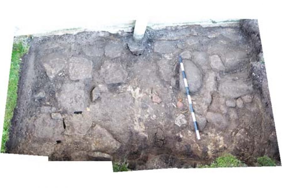 En arkeologisk utgrävning visar en rektangulär yta med stenar och jord. En mätsticka ligger diagonalt över området för att ge storleksperspektiv. Området verkar vara resterna av en gammal struktur, möjligen en kyrka.