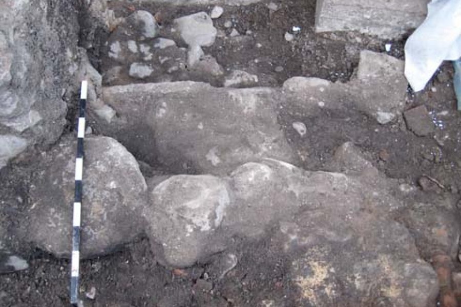 Stenstrukturer grävs fram i arkeologiskt utgrävningsområde i Gamla Stan, omgivna av jord och stenar. En skala med svart-vita markeringar ligger intill för att visa storlek och proportioner.