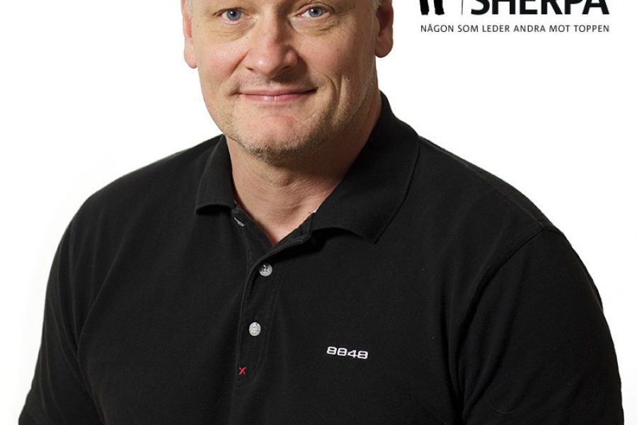 Man i svart pikétröja står framför vit bakgrund. Texten bredvid honom lyder "Årets Sherpa" och "Någon som leder andra mot toppen" med en siluett av en bergsklättrare och berg. Han ler vänligt.