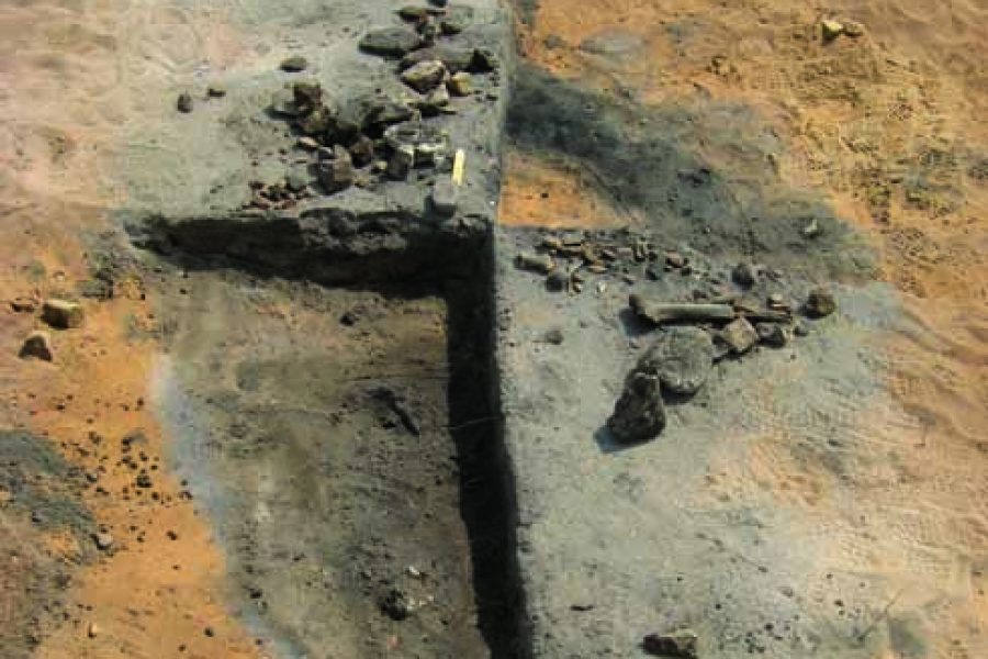 Ett arkeologiskt utgrävningsområde visar en rektangulär jordformation med en tydlig vallgrav. Stenar och fragment är utspridda över ytan. Marken är sandfärgad, vilket tyder på en plats med historisk betydelse.