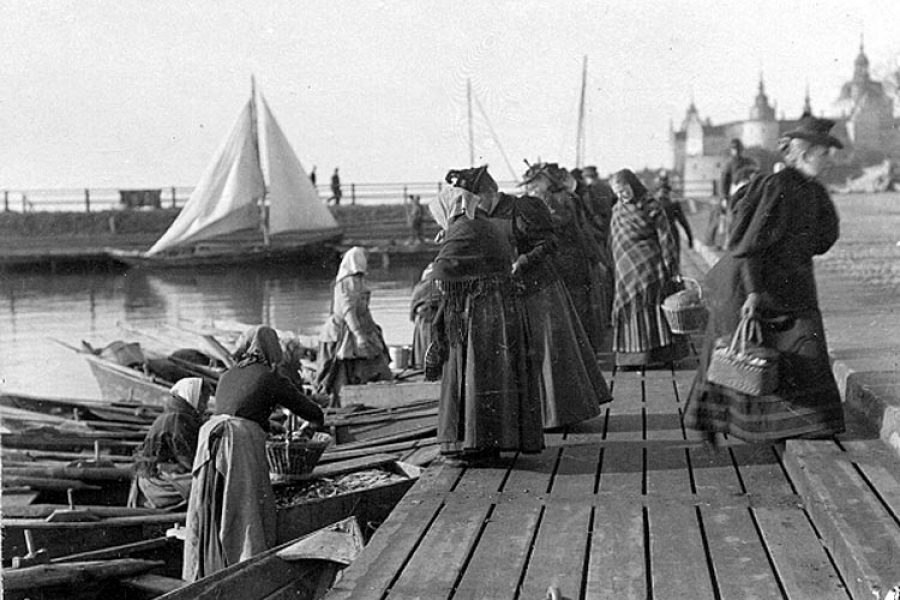 Kvinnor i traditionella kläder handlar fisk från båtar vid en träbrygga i en historisk hamn. En segelbåt syns i bakgrunden. Byggnader med tinnar och torn markerar horisonten. Soligt väder bidrar till en livlig atmosfär.