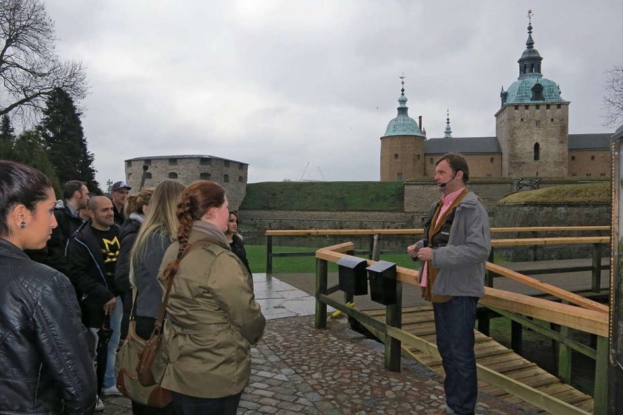 En guide håller en föreläsning utomhus framför en grupp människor vid ett historiskt slott med flera torn. Deltagarna lyssnar uppmärksamt. Slottet ligger i bakgrunden med molnig himmel ovanför.