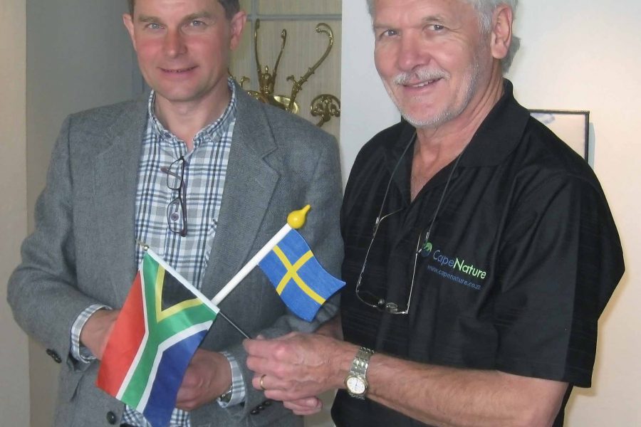 Två män står inomhus och håller i flaggor; den ena den sydafrikanska och den andra den svenska. Mannen till vänster bär en grå kavaj, medan mannen till höger har en svart skjorta med texten "CapeNature". De ser glada ut.