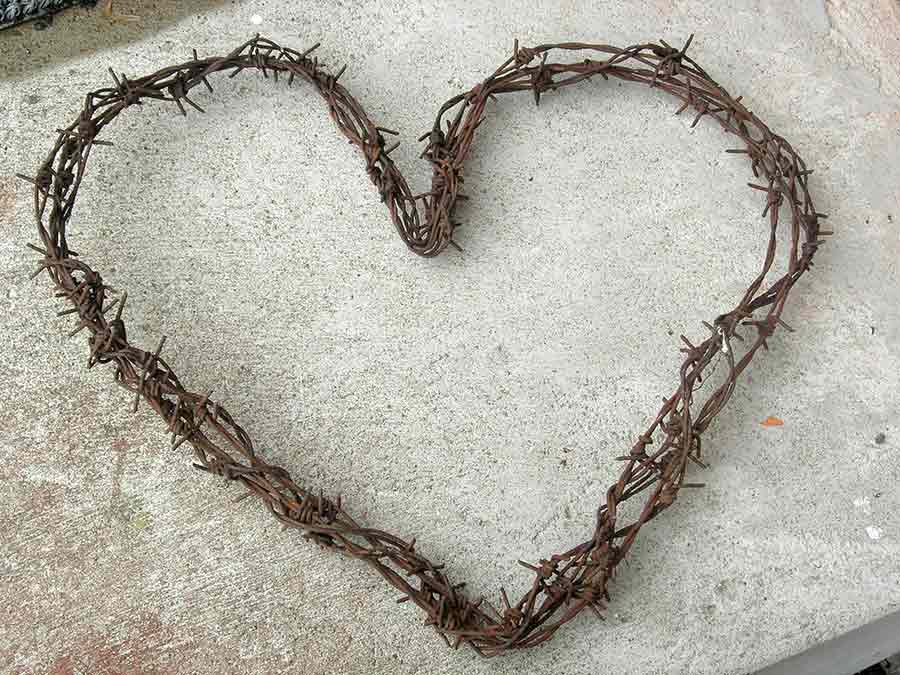 Ett hjärta format av rostig taggtråd ligger på en grov, ljusgrå betongyta. Taggtråden är tät med vassa taggar, vilket skapar en kontrast mellan mjuk form och hårt material, symboliserande konflikt och skönhet.