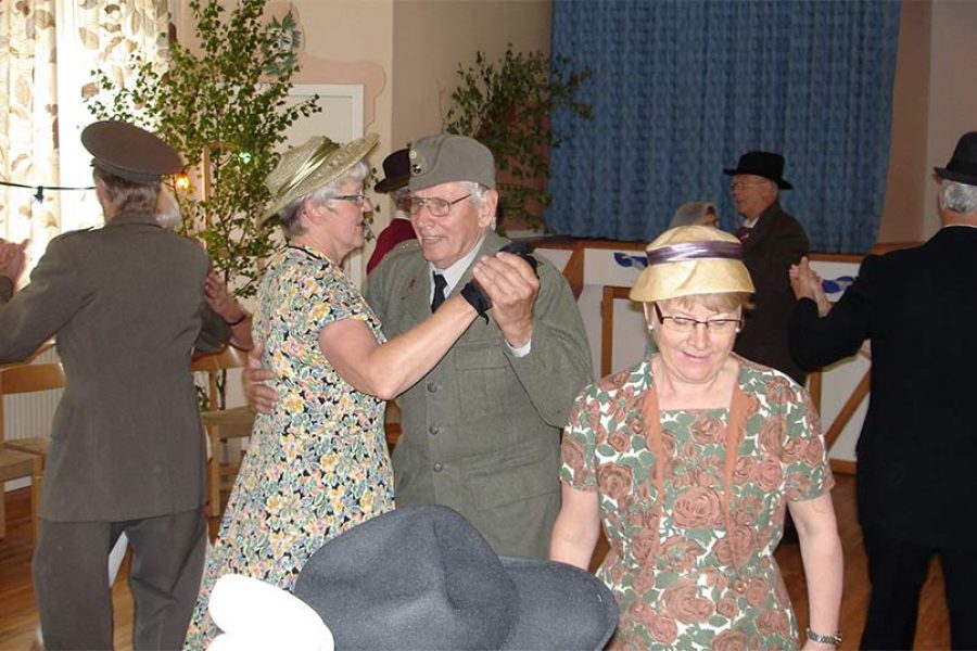 Äldre par dansar glatt i en dekorativt utsmyckad sal med trämöbler och blommig tapet. Kvinnorna bär klänningar och hattar, medan männen har kostymer. I bakgrunden syns fler personer dansa i par.