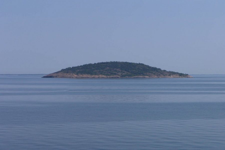 En ö med skogtäckt terräng sträcker sig lågt över det stilla vattnet under en klarblå himmel. Omges av hav, kan en skogsbeklädd kulle ses mitt i bilden som antyder ett fridfullt och avlägset område.