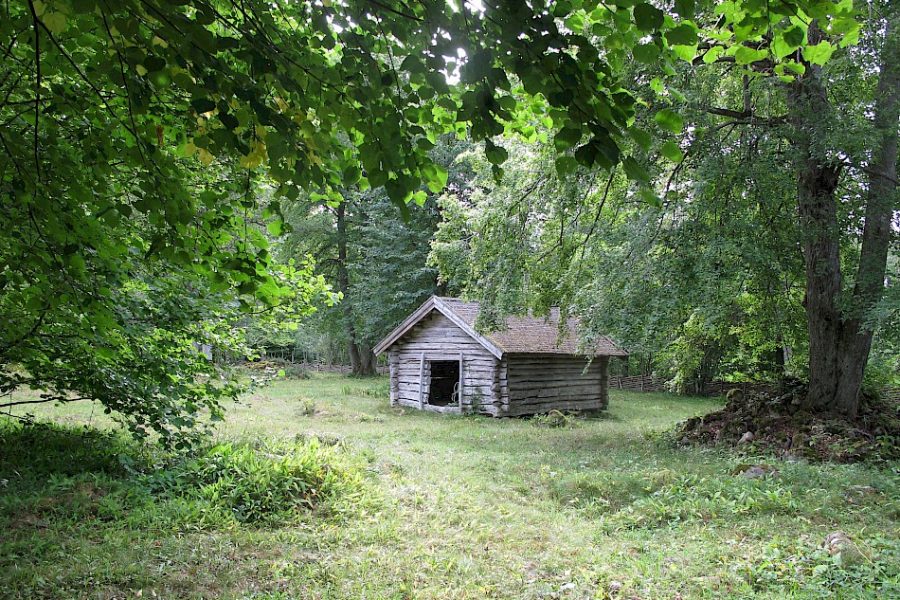 En träbyggnad med trasigt tak står på en grönskande äng omgiven av täta lövträd. Skogen skapar en lugn och stillsam atmosfär runt det lilla huset, som verkar övergivet och historiskt.