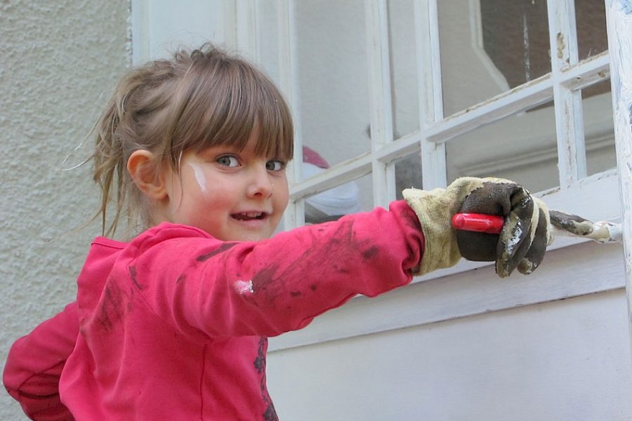 En flicka i rosa tröja och handskar målar en vit fönsterkarm med en pensel. Hennes kläder och handskar är smutsiga av färg, och hon ler medan hon tittar mot kameran.