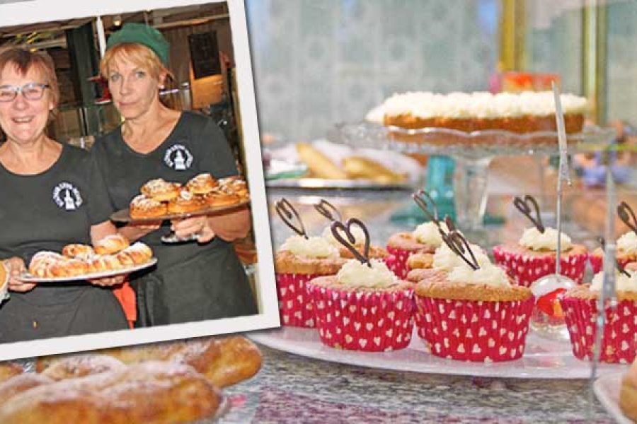 Tre personer i köksuniformer, med gröna hattar, håller brickor med bakverk, inklusive bullar och en blåbärspaj. I förgrunden syns en monter med cupcakes i röda formar. Scenen antyder en kafénominering 2014.