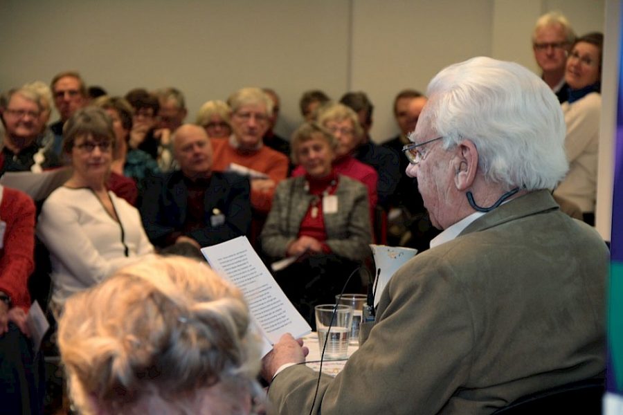 En äldre man med glasögon läser från ett papper framför en intresserad publik. Åhörarna sitter tätt, några med namnlappar, och lyssnar noga i en konferensliknande miljö. I förgrunden syns en kvinna.