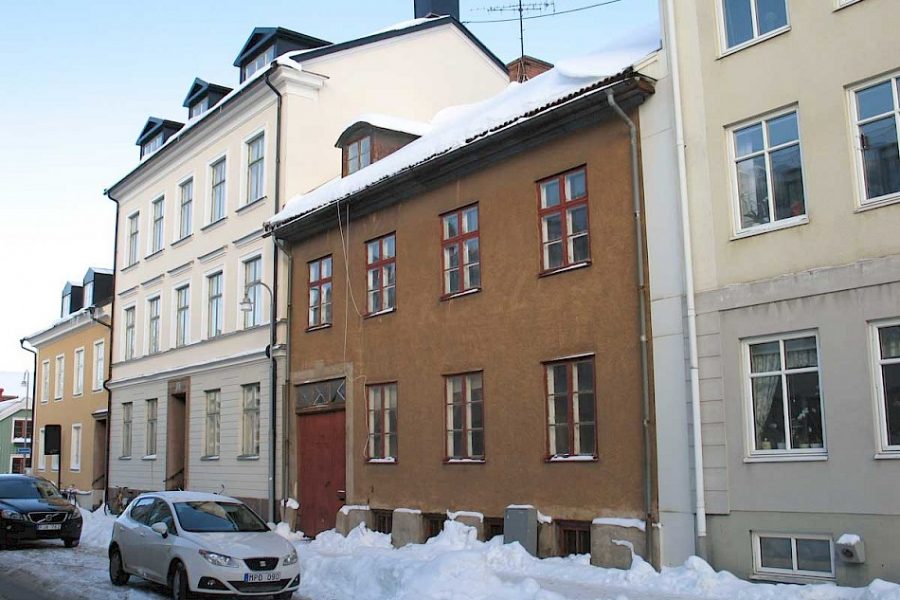 En vintrig gata kantad av äldre byggnader med snötäckta tak. Två parkerade bilar syns framför en brun tvåvåningsbyggnad. Klar blå himmel och smältande snö ger en känsla av kylig vinterdag i stadsmiljö.