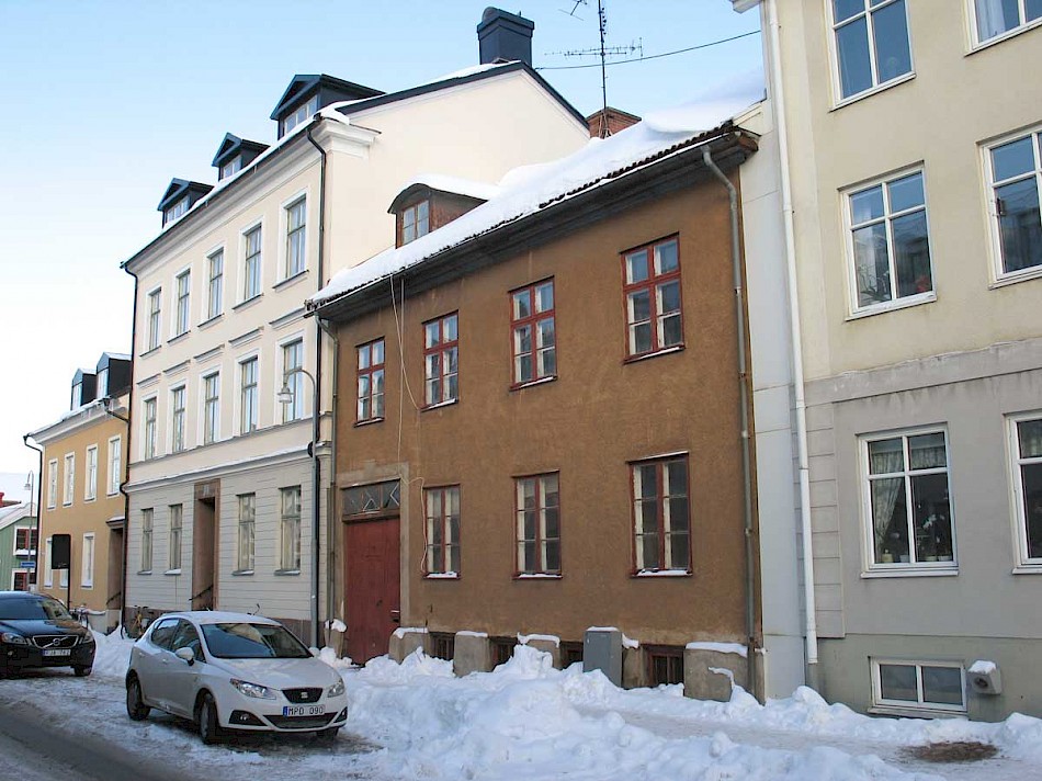 En vintrig gata kantad av äldre byggnader med snötäckta tak. Två parkerade bilar syns framför en brun tvåvåningsbyggnad. Klar blå himmel och smältande snö ger en känsla av kylig vinterdag i stadsmiljö.