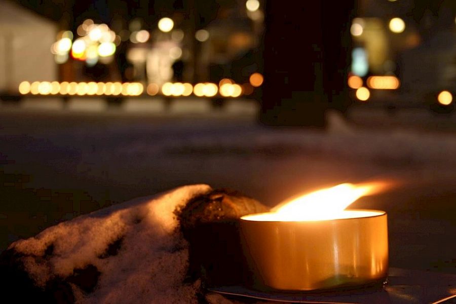 Ett ensamt ljus brinner på en snötäckt yta i förgrunden, medan en rad av suddiga ljus lyser upp bakgrunden i en nattlig miljö, vilket skapar en stämningsfull och minnesvärd atmosfär.