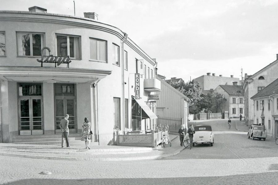Saga biograf i Oskarshamn med neonskylt, bredvid konditori. Två personer står framför ingången medan cyklister och bilar passerar på gatan. Omgiven av äldre byggnader och träd i bakgrunden. Svartvitt foto.
