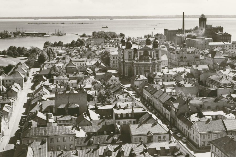 Svartvit flygbild över en historisk stad med många trähus och några större byggnader. I bakgrunden syns en fabrik och vatten. Landskapet känns idylliskt och tidstypiskt med stadens arkitektur och närheten till kusten.