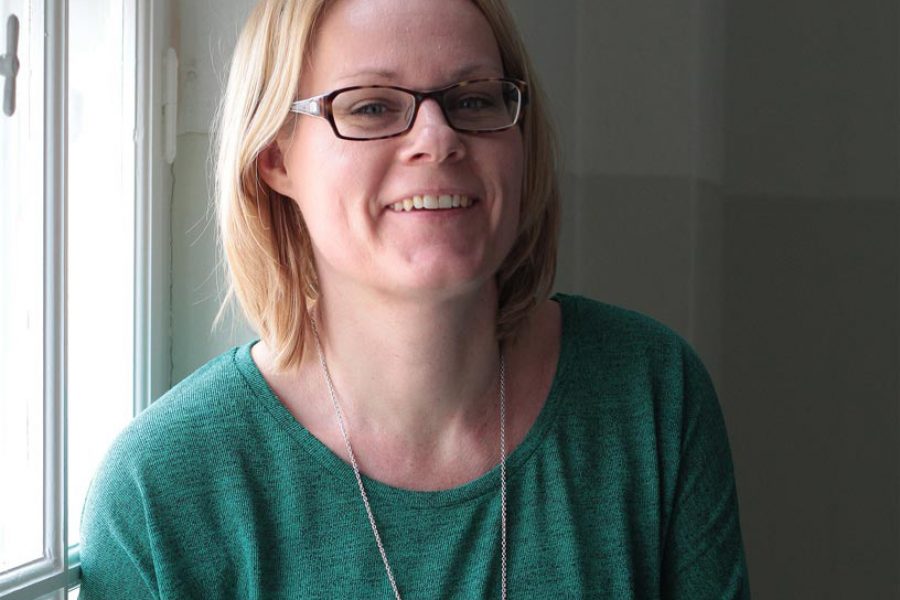 En person med glasögon och ljust hår ler i naturligt ljus, stående nära ett fönster. Personen bär en grön tröja och ett långt halsband. Bakgrunden är en ljus vägg som accentuerar det mjuka ljuset.