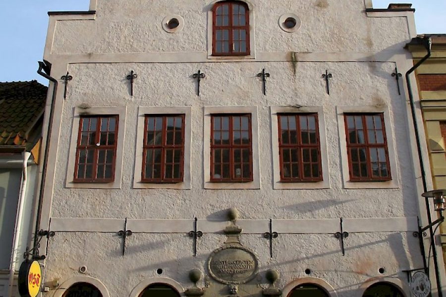 Gammal byggnad med vit fasad och röda fönster i tre våningar. Överst en trapetsformad gavel. Bottenvåningen har tre välvda fönster och en dörr med dekorativa stendetaljer. Skyltar och produktvisningar finns i fönstren.