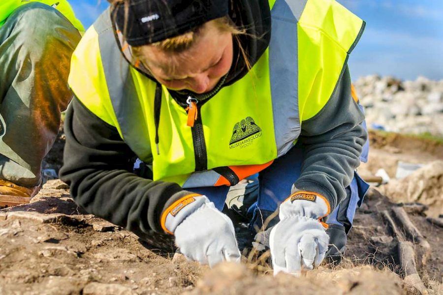 En person i reflexväst och handskar gräver försiktigt i jorden, troligen på en arkeologisk utgrävning av ett skelett. Solig utomhusmiljö med stenar i bakgrunden. Text på västen: “Mussarkeologi Sydost”.