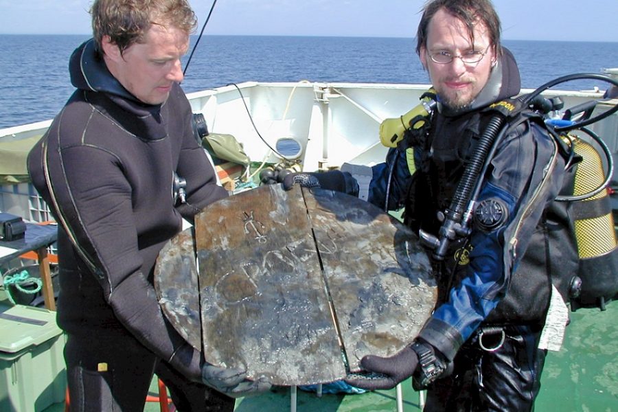 Två dykare i våtdräkter håller en stor rund skiva av trä med inskriptioner, stående på ett fartygsdäck med havet i bakgrunden. Solen skiner och utrustning syns i bakgrunden.