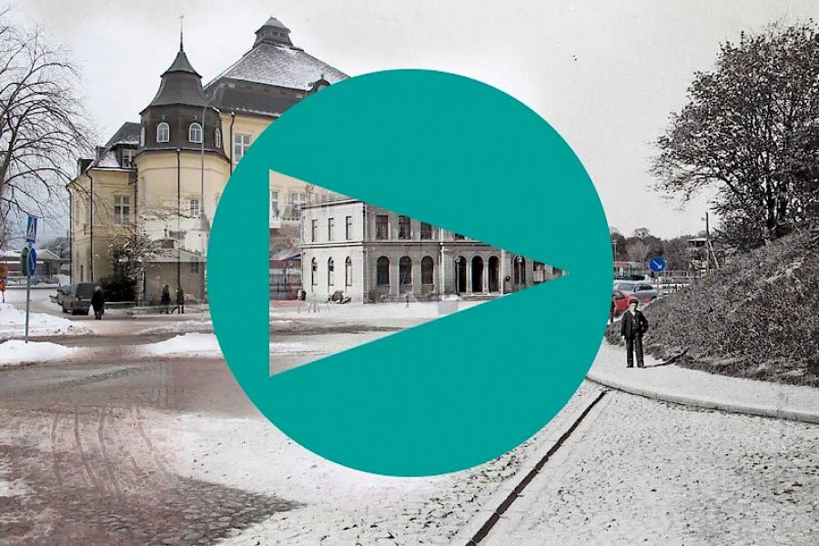 I don't have the capability to view images, but here is a possible alt text based on the file name and description:

Stort tealfärgat spelsymbol över en vinterlandskapsscen i Kalmar. Snö täcker marken framför äldre byggnader och ett avlägset par som promenerar. Byggnaderna har historisk arkitektur och omges av träd.