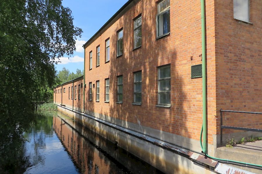 Tegelfasad reflekteras i en lugn vattenspegel, kompletterad av omgivande grönska och klar blå himmel. Byggnadens fönster kantar kanalen, vilket skapar en harmonisk och fridfull miljö. Lummiga träd ramar in scenen.