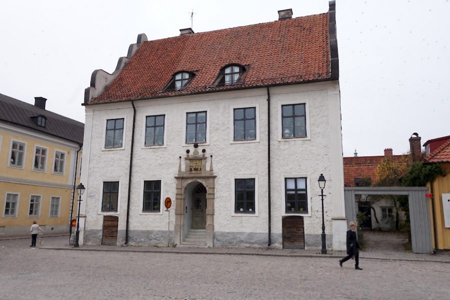 Ett vitputsat stenhus med röd tegeltak står vid en kullerstensgata. Två personer går förbi, en på varje sida. Detaljerad dörröppning i sten, sex stora fönster på fasaden. Brevidliggande byggnader ses runtom.