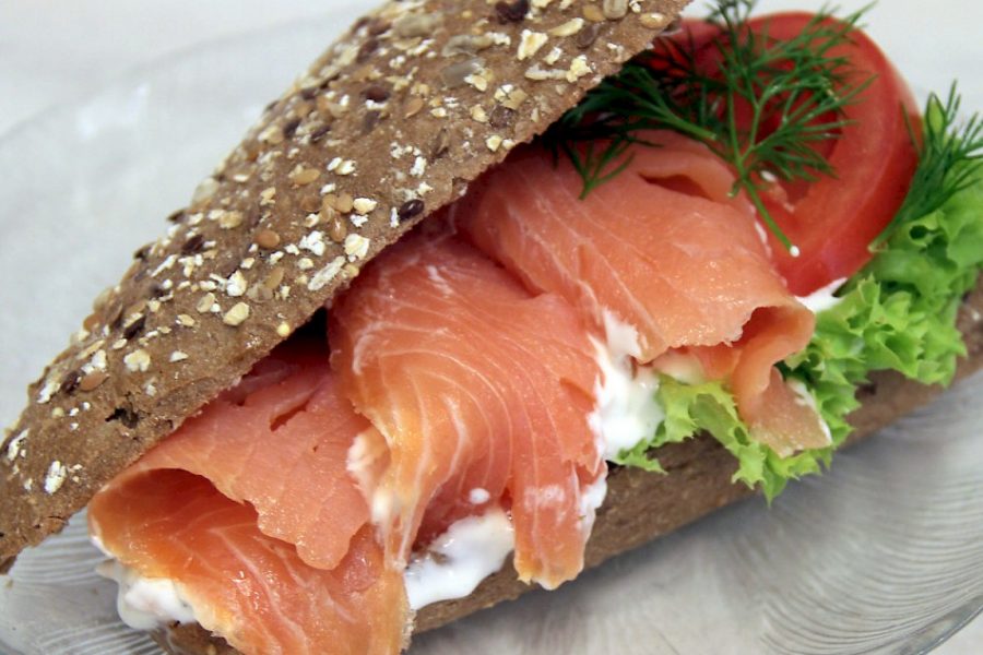 Fullkornsbröd fyllt med skivad lax, sallad, tomat och dill på en tallrik. En klick krämig sås syns mellan laxen och brödet. Pumpafrön och havreflingor toppar brödets yta för extra textur.