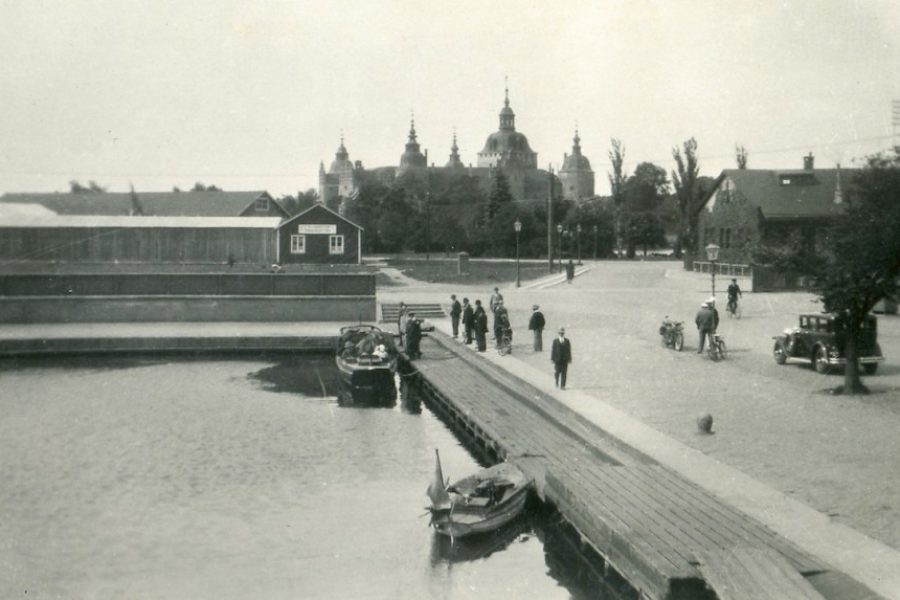 En småbåt ligger förtöjd vid en brygga i en hamn, omgiven av människor som promenerar och cyklar. I bakgrunden syns ett slott med flera torn. Bilden verkar vara från omkring 1930.