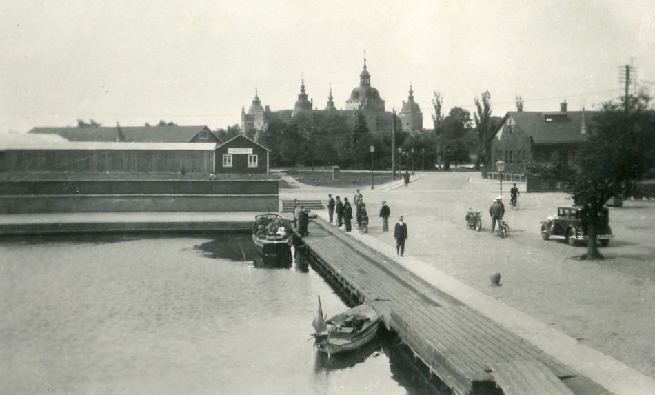En småbåt ligger förtöjd vid en brygga i en hamn, omgiven av människor som promenerar och cyklar. I bakgrunden syns ett slott med flera torn. Bilden verkar vara från omkring 1930.
