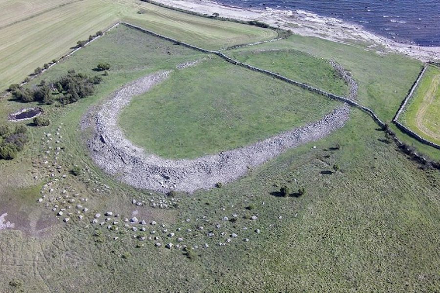 Flygvy över Ales stenar, en megalitisk stenformation i form av ett skepp, belägen på en grässlätt nära kusten. Omgivande land består av fält och små buskage. Havet syns i bildens övre del.