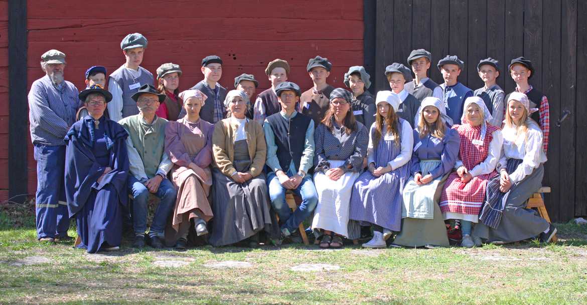 En grupp människor, klädda i tidstypiska kläder från början av 1900-talet, poserar framför en röd och svart byggnad. De står och sitter i två rader på en gräsmatta i starkt solljus.