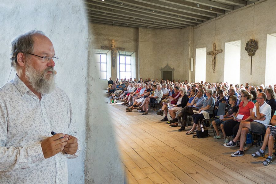 Man står vid fönster och håller tal inför stor publik i en rustik sal. Deltagarna sitter på stolar i rader, lyssnande, med synligt intresse. Ljus strömmar in genom fönster, dekorationer pryder väggarna.