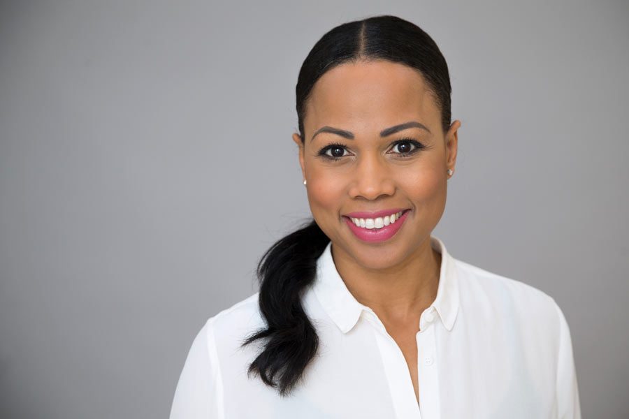 Kvinna med svart hår i hästsvans, klädd i vit skjorta, ler mot kameran. Bakgrunden är grå och personen verkar avslappnad och positiv. Hennes leende och ljus läppstift ger ett vänligt intryck.