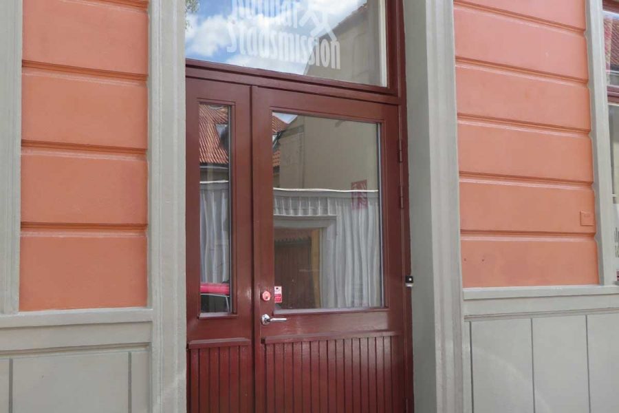 Dörr till Kalmar Stadsmission i en byggnad med orange fasad och gråa dörrkarmar. Fönstret ovanför dörren har texten "Kalmar Stadsmission". En blomkruka med gula och gröna växter står vid sidan av dörren.