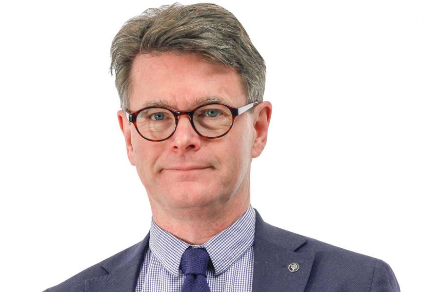 Man med kort hår bär glasögon och en mörkblå kavaj över en randig skjorta med slips. Står framför en vit bakgrund. Känns formellt klädd, som för en professionell porträttbild.