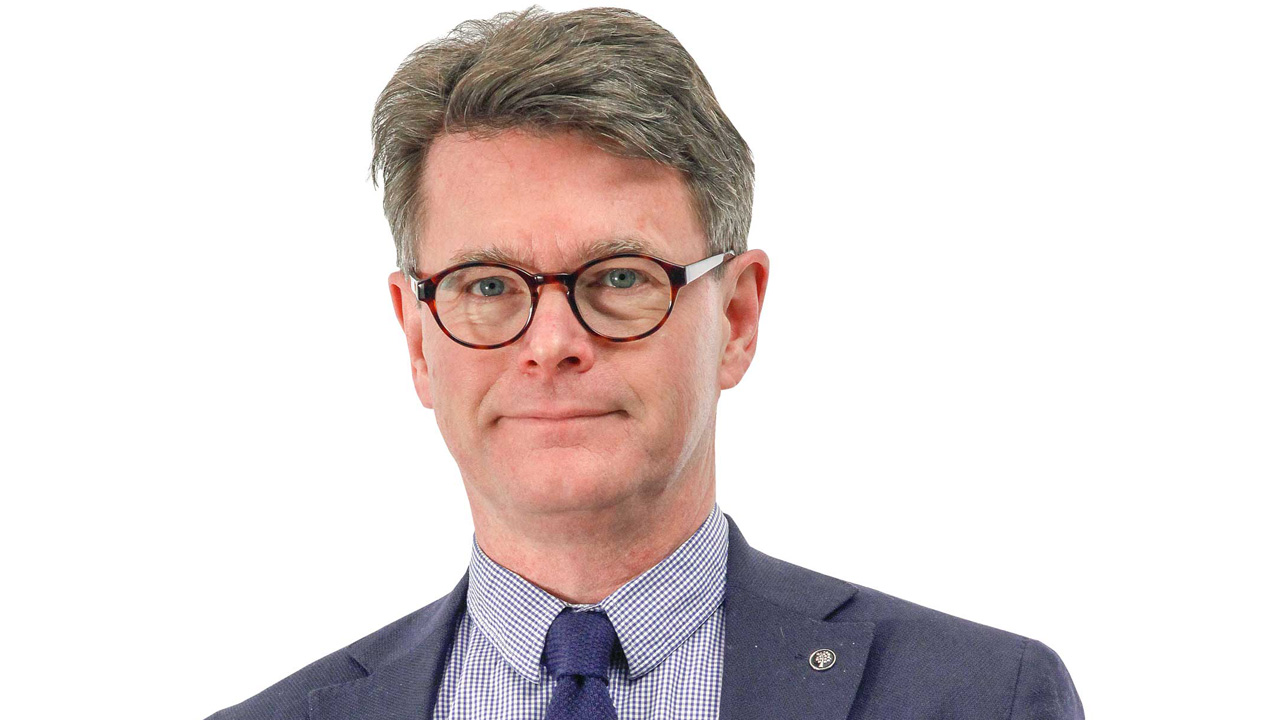 Man med kort hår bär glasögon och en mörkblå kavaj över en randig skjorta med slips. Står framför en vit bakgrund. Känns formellt klädd, som för en professionell porträttbild.