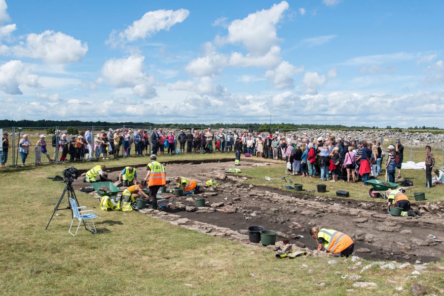 Arkeologer i reflexvästar gräver i marken vid en utgrävning på Sandby borg. En stor publik står bakom avspärrningar och observerar. Himlen är molnig och landskapet runt omkring är öppet och naturskönt.