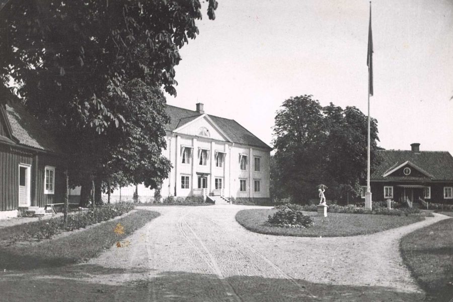 Stort tvåvåningshus i vit panel med klassiska kolonner, omgivet av lummiga träd. I förgrunden finns en grusad uppfart med en staty på en gräsmatta och en flaggstång. Mindre hus på var sida.