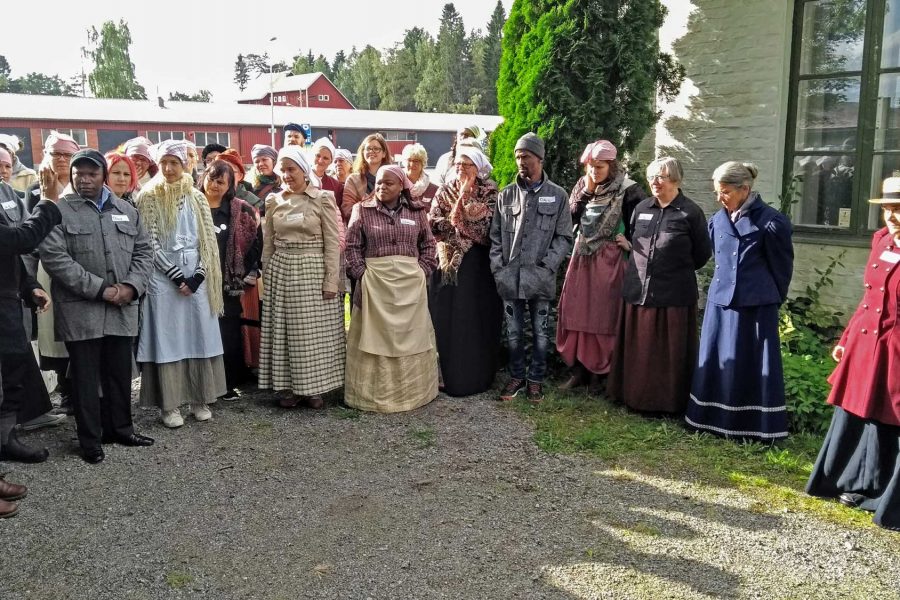 En grupp människor klädda i tidstypiska kläder från tidigt 1900-tal står i en halvcirkel utomhus. De lyssnar på en man som gestikulerar. Byggnader och träd finns i bakgrunden.
