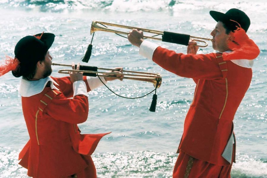 Trumpetblåsare i röd uniform spelar vid havet. De bär svarta hattar med röda fjädrar och håller gyllene trumpeter. Solen glittrar på vattnet i bakgrunden, vilket skapar en livlig atmosfär.