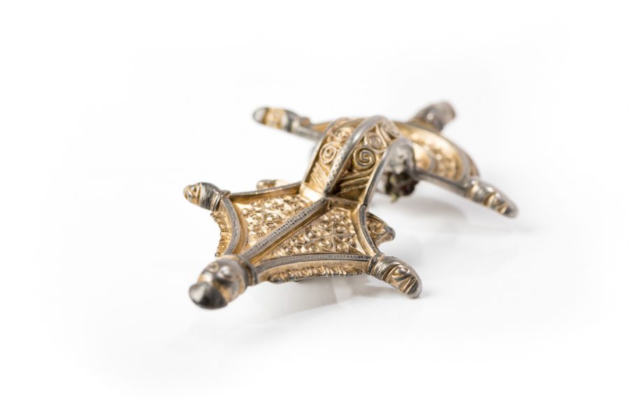 Dekorativt metallspänne av guld och silver, vilande på vit bakgrund. Spännet har intrikata spiralmönster och upphöjda detaljer som skapar en tredimensionell effekt. Det framhäver konstnärligt hantverk från en tidigare era.