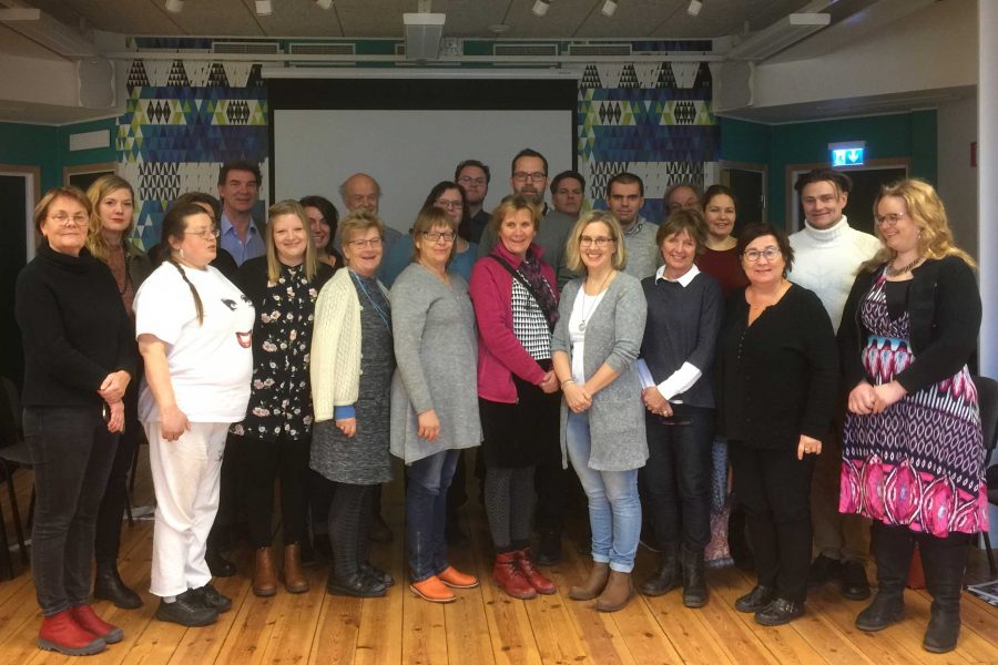 En grupp av cirka 20 personer står tillsammans i ett rum med trägolv och färgglatt mönstrade väggar. De flesta ler och verkar delta i ett möte eller en sammankomst, kanske i en kulturell eller utbildningsmiljö.