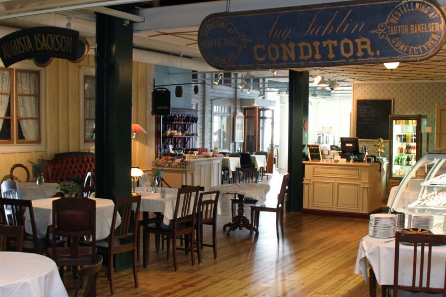 En inbjudande restauranginteriör med träväggar och trägolv. Flera runda bord är dukade med vita dukar och omgivna av stolar. En disk i bakgrunden visar bakverk och en skylt med texten "Augusta Isackson".