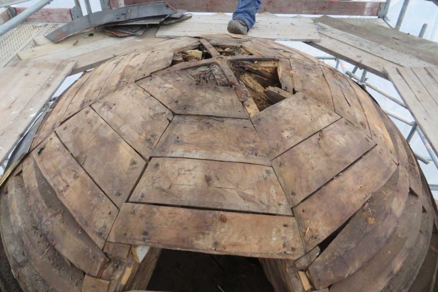 Trätak med synliga skador och hål i mitten, omgiven av en byggnadsställning. En person står nära och inspekterar. Träplankorna är slitna och täckta av smuts, vilket indikerar behov av restaurering.