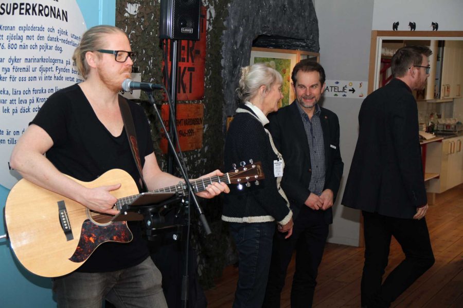 Man spelar akustisk gitarr och sjunger framför mikrofonen. Två personer samtalar bredvid, och en tredje går bortåt. Konst och text om "Superkronan" syns på väggen. Musikerens mikrofon är ansluten till högtalare.