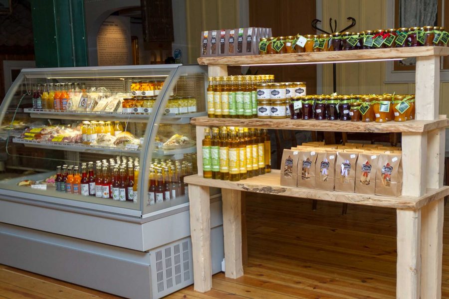 Kylskåp med glasruta full med juiceflaskor och påsar med charkvaror. Trähyllor bredvid visar flaskor med olivolja, syltburkar och bruna papperspåsar. Trägolv och museimiljö indikerar lokal plats i Kalmar läns museum.