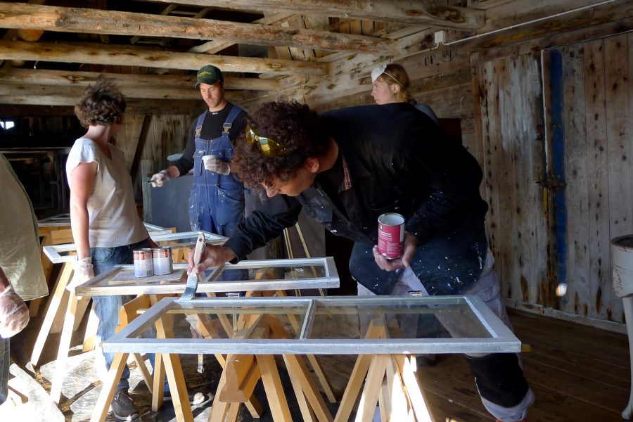 Personer deltar i en workshop för fönsterrenovering i en rustik träbyggnad. En person målar fönsterkarmar med en pensel medan andra observerar och diskuterar processen. Byggnaden har synliga träbjälkar och gamla dörrar.