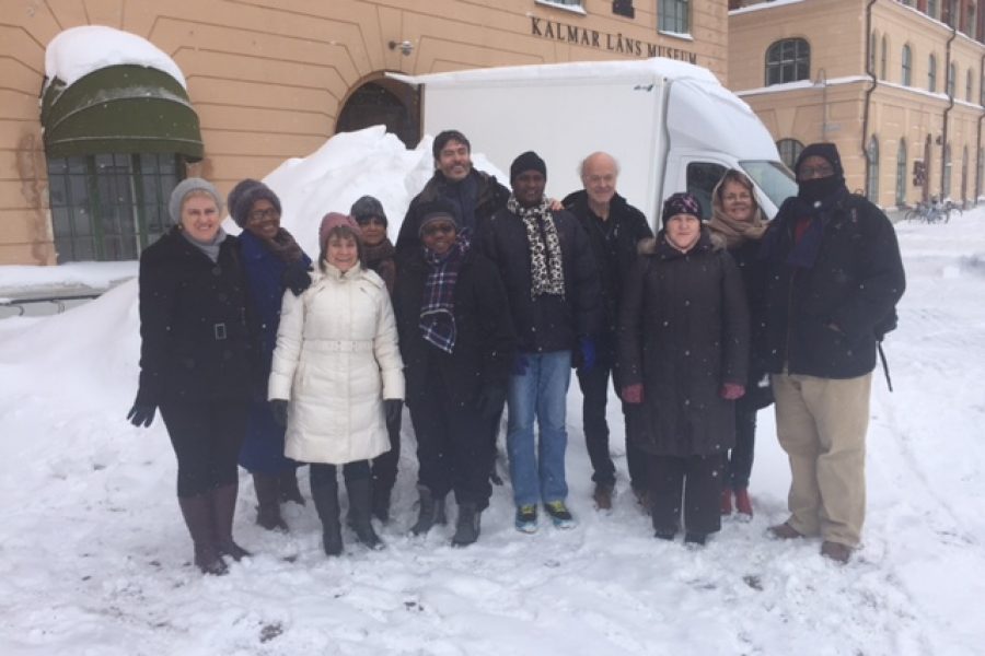 En grupp på tolv personer står framför en snötäckt byggnad med skylten "Kalmar Läns Museum". De bär vinterkläder såsom jackor och halsdukar. Snö faller och täcker marken runt dem.