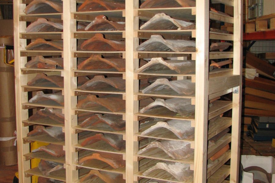 Träställ med rader av tegelpannor i varierande nyanser av brunt och grått, ordnade ordentligt på hyllor. Stället står inomhus på ett trägolv inom Kalmar läns museums samlingar, vilket antyder en arkivmiljö.